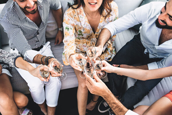 Cheerful friends sitting in the terrace garden couch having fun toasting by clinking champagne flute glasses - happiness lifestyle concept - high angle view